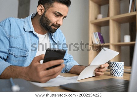 Young indian businessman holding phone reading bank receipt calculating taxes, ethnic man using smartphone mobile application checking bill document, managing money finances, loan expenses.