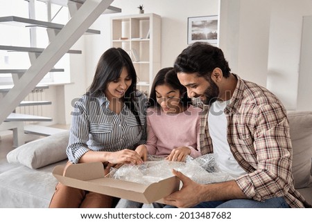 Excited indian family with child daughter unpack parcel at home. Happy parents and teen kid daughter open postal box receive present gift in online shopping delivery package sitting on sofa together.
