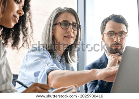 Mature older ceo businesswoman mentor in glasses negotiating growth business plan with diverse executive managers at boardroom meeting table using laptop. Multicultural team work together in office.