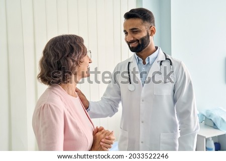 Happy young Indian doctor therapist in white coat has appointment consulting supporting putting hand on shoulder of older senior female patient in modern clinic hospital. Medical healthcare concept.