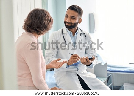 Indian male doctor consulting senior old patient filling form at consultation. Professional physician wearing white coat talking to mature woman signing medical paper at appointment visit in clinic.