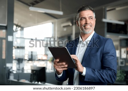 Happy middle aged business man ceo wearing suit standing in office using digital tablet. Smiling mature businessman professional executive manager looking away thinking working on tech device. 商業照片 © 