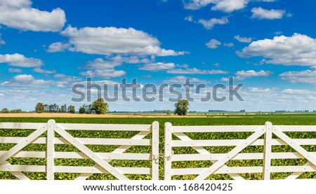 A white picket fence gate in front of a beautiful farmland on a sunny day