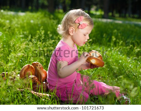The little blonde beautiful girl sitting on a green lawn in the park and eat a muffin.