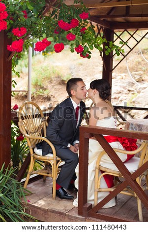 Young couple in love the bride and groom posing in the arbor under red roses, wedding day in the summer. Enjoying a moment of happiness and love.