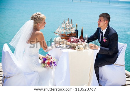 Beautiful couple in love the bride and groom posing on the bridge at the sea at the wedding table decorated with exotic bottles and wooden model ship. Enjoy a moment of happiness and love.
