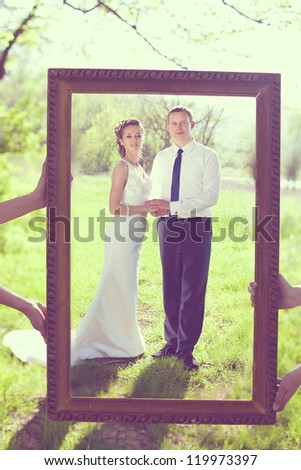 Portrait of the bride and groom are posing with the big frame in the park.