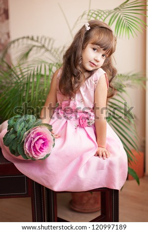 Beautiful little girl model view in a luxury pink dress and flower posing at home on the table.