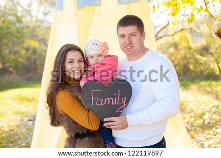 Happy young family mom dad and little baby posing on background of the tent and trees. Held in the hand plate in form of heart with the words FAMILY.