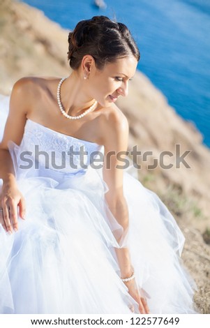 Beautiful young bride dressed in luxurious white dress posing sitting on a stone on cliff against the blue sea