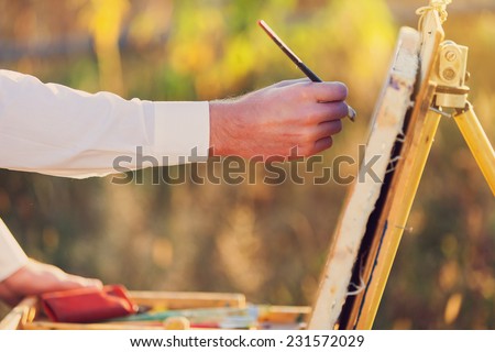 Box easel with canvas, oil paint, palette, and tools. Hand of painter sunset.