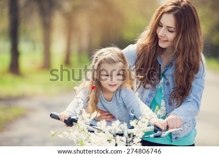 Beautiful young mother teaching her daughter to ride a bicycle. Both smiling, summer park in background, active family concept