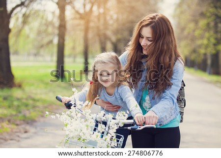 Beautiful young mother teaching her daughter to ride a bicycle. Both smiling, summer park in background, active family concept