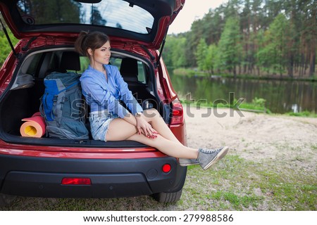 Happy young woman tourist travel on a red car to summer forest. Holiday trip to lake