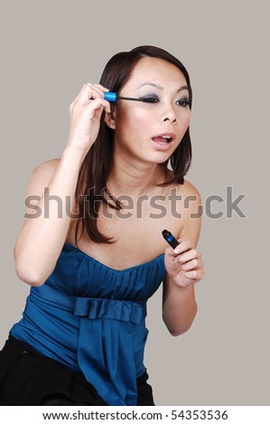 A beautiful brunette Chinese woman, doing her makeup  and fixing her eyelashes, for light gray background.