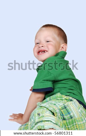 An young toddler boy has fun playing on the floor in the studio, shooing his missing tooth, in a green outfit on light blue background.