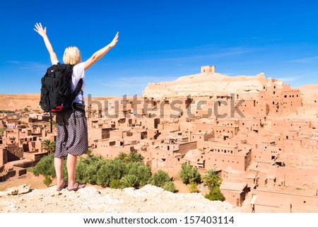 Adventurous woman arms reised in front of Ait Benhaddou, fortified city, kasbah or ksar, along the former caravan route between Sahara and Marrakesh in present day Morocco.