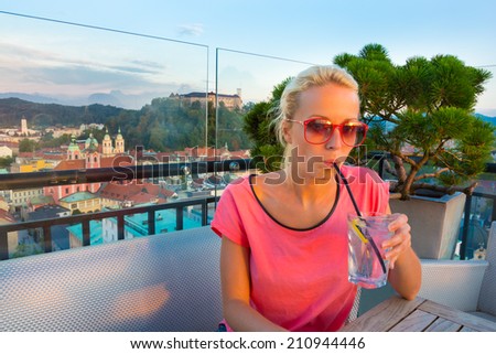 Female tourist enjoying refreshment drink in Neboticnik rooftop bar with panoramic view of Slovenian capital Ljubljana at sunset.