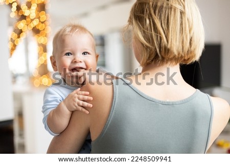 Foto Bild Blonde Mutter hält ihr bezauberndes fröhliches kleines Baby im Arm. Nettes Kleinkind Kind in die Kamera schauen und lächelnd zu Hause