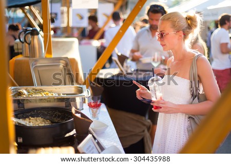 Beautiful blonde caucasian lady wearing white summer dress buying freshly prepared mealat a local food market. Urban international kitchen event taking place in Ljubljana, Slovenia, in summertime.