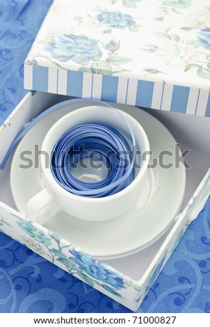 Gift box with white cup saucer and a blue ribbon in it on blue background