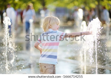 Little boy plays in the square between the water jets in the fountain at sunny summer day. Active summer leisure for kids in the city.