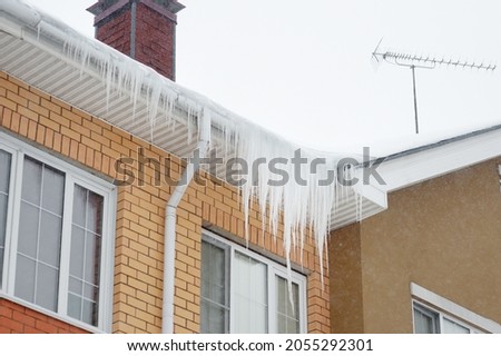 Big icicles on the roof of a townhouse on a snowy winter day among thaw. Cleaning the roofing from snow and icicles.