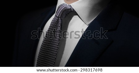 A dramatic close up shot of a man dressed in an expensive formal suit. The jacket is dark navy with a matching, knitted, dark navy and white tie and an intricately detailed white buttoned up shirt.