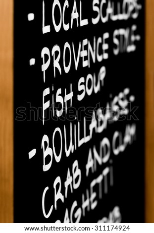 Example of a typical menu drawn on a black chalkboard. Featured seafood dishes include: local scallops, provence style fish soup and also bouillabaisse crab and clam spaghetti. Shallow depth of field.
