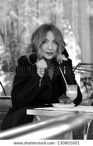 Black and white portrait of a happy young woman sitting at a table in a cafe drinking a cappuccino and eating cake