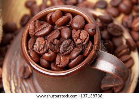 Delicious dark brown coffee beans in small ceramic coffee cup over light brown wooden background