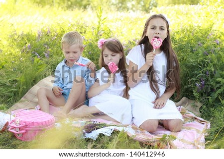 Three children sitting on the rug in the grass and eat lollipops outdoors
