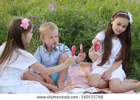 Three children sitting on the rug in the grass and eat lollipops