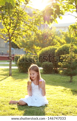 young girl with a sweet smile in white dress sitting on grass and eating ice cream on a hot sunny day