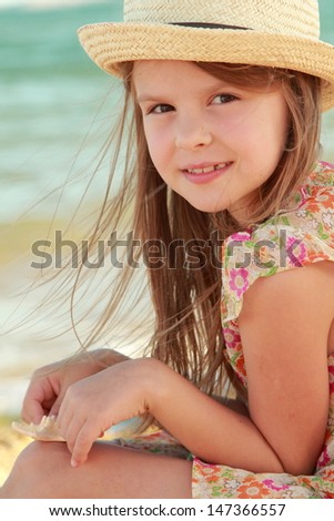 Charming joyful little girl in a summer dress dreaming and smiling at the beach on a summer day/Lovely girl with a beautiful smile playing barefoot in the water on the beach
