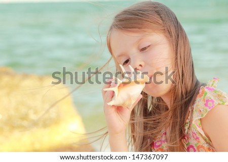 Charming joyful little girl in a summer dress dreaming and smiling at the beach on a summer day/Lovely girl with a beautiful smile playing barefoot in the water on the beach
