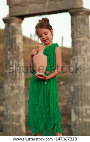 Beautiful young Greek goddess in emerald green vintage dress holding of an ancient amphora on the background of the ruins of the ancient city