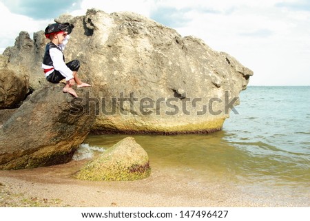 Angry little boy dressed as a pirate is sitting on a large rock in the sea and waiting for the ship