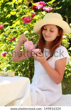 Happy little girl in cute hat playing with children\'s dishes in the tea party outdoors