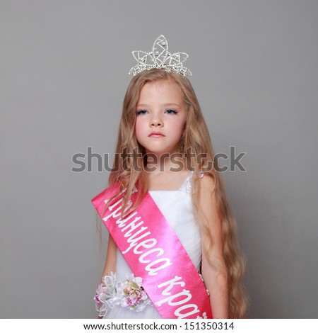 Charming little blonde in a beautiful white dress and a crown won the competition little princesses/girl in a white dress with a red ribbon and inscription in Ukrainian \