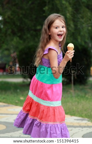 Happy cute girl in a bright dress is eating strawberry ice cream on a background of the park on a sunny summer day/Child eats ice cream in the park