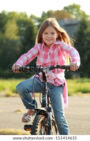 Charming young girl in blue jeans rides a bicycle in a park on a sunny day/Caucasian smiling child playing sports on a bicycle in a park outdoors
