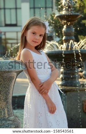 Lovely little girl in white dress sitting on parapet near the fountain in the summer hottest day/Portrait of beautiful girl near the fountain