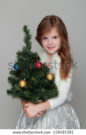 Charming cheerful little girl in a shiny dress holding a small Christmas tree with balls on a gray background for the New Year