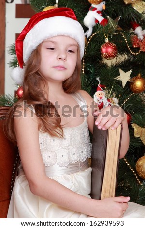 Attractive smiley little girl holding dark brown vintage book over Christmas decoration on Holiday theme/Cheerful young girl wearing santa\'s hat and holding big ancient book