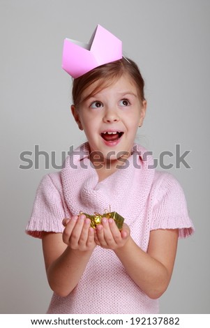 beautiful Caucasian girl with a charming smile in a knitted dress with a pink crown holding a gift on a gray background/Beauty little princess with pink tiara