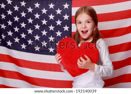Image of a beautiful cheerful little girl with a sweet smile, holding a big red heart on the background of the American flag on Holiday