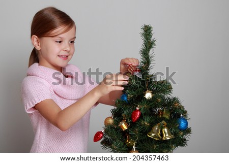 Portrait of a cute child in a knitted dress decorates a Christmas tree on a gray background on Holiday/Smiling girl near a Christmas tree