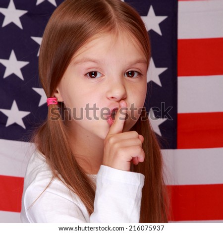 Happy smiling little girl put her finger to her lips as a sign of silence on the background of a large U.S. flag