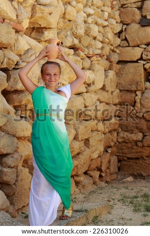 Happy cheerful little girl in a Greek tunic as a Greek goddess holding a vintage amphora on the background of the archaeological site of the ancient city of Nymphaeum in the village Geroevskoe